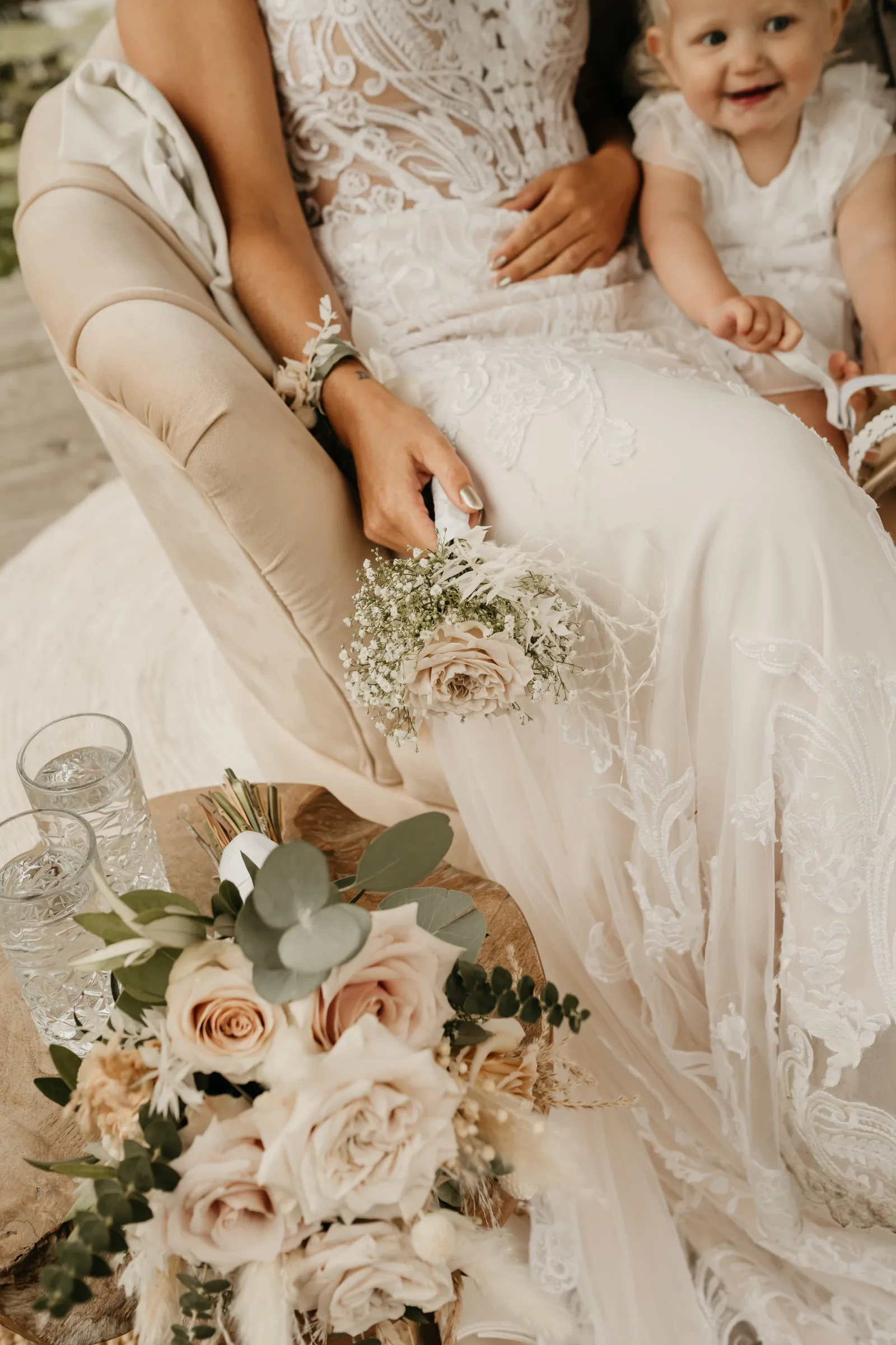 Een bruid zit in een crème fluwelen fauteuil, in beeld vanaf haar schouders: een kanten bovenstuk met bloemapplicatie, een corsage van droogbloemen om haar pols en een klein boeketje blush rozen in haar handen. Rechts naast haar een lachend meisje in een wit kanten jurkje met bungelende benen.