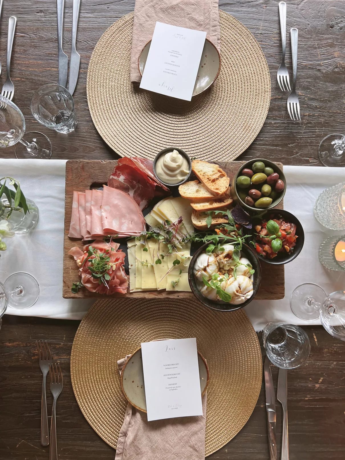 Een deelplank van bovenaf gefotografeerd: dun gesneden ham, kaasblokjes, olijven, tomaatjes, brood en dipjes in schaaltjes, op een witte loper met ronde gevlochten placemats, bestek en een menukaartje ernaast.
