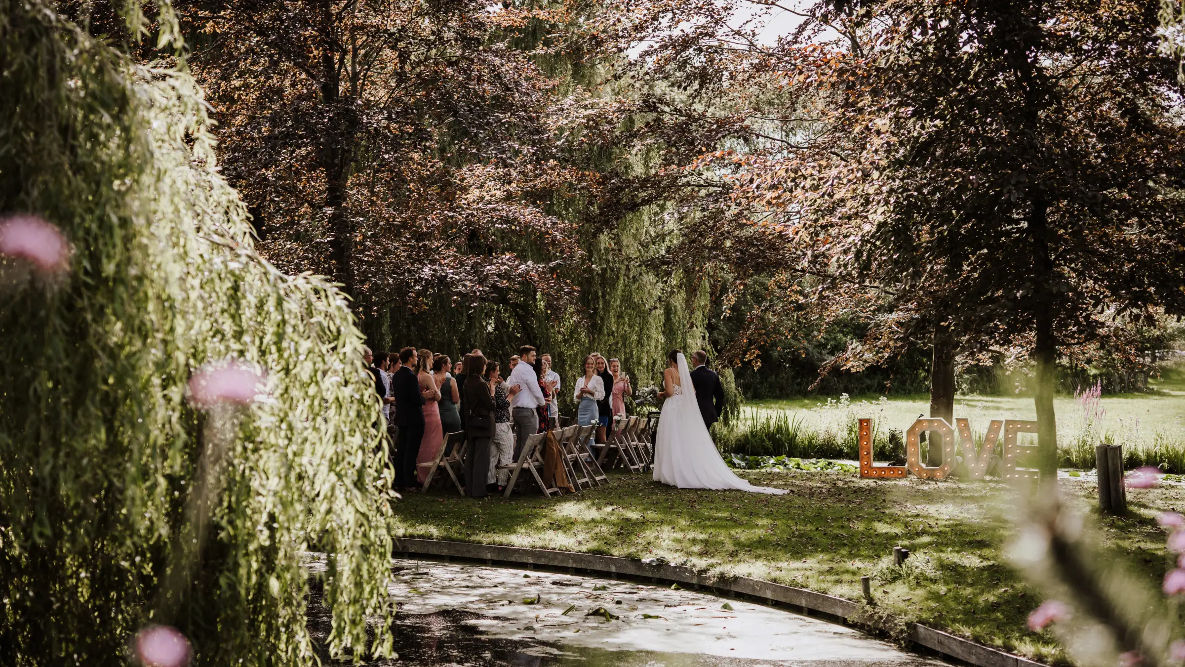 De ceremonie op het eiland gezien door de hangende takken van een treurwilg: gasten staan en zitten langs het water, de bruid loopt met een lange sleep over het gras, en rechts aan de waterkant staan houten LOVE-letters. Op de voorgrond hangen roze bloemen onscherp in beeld.
