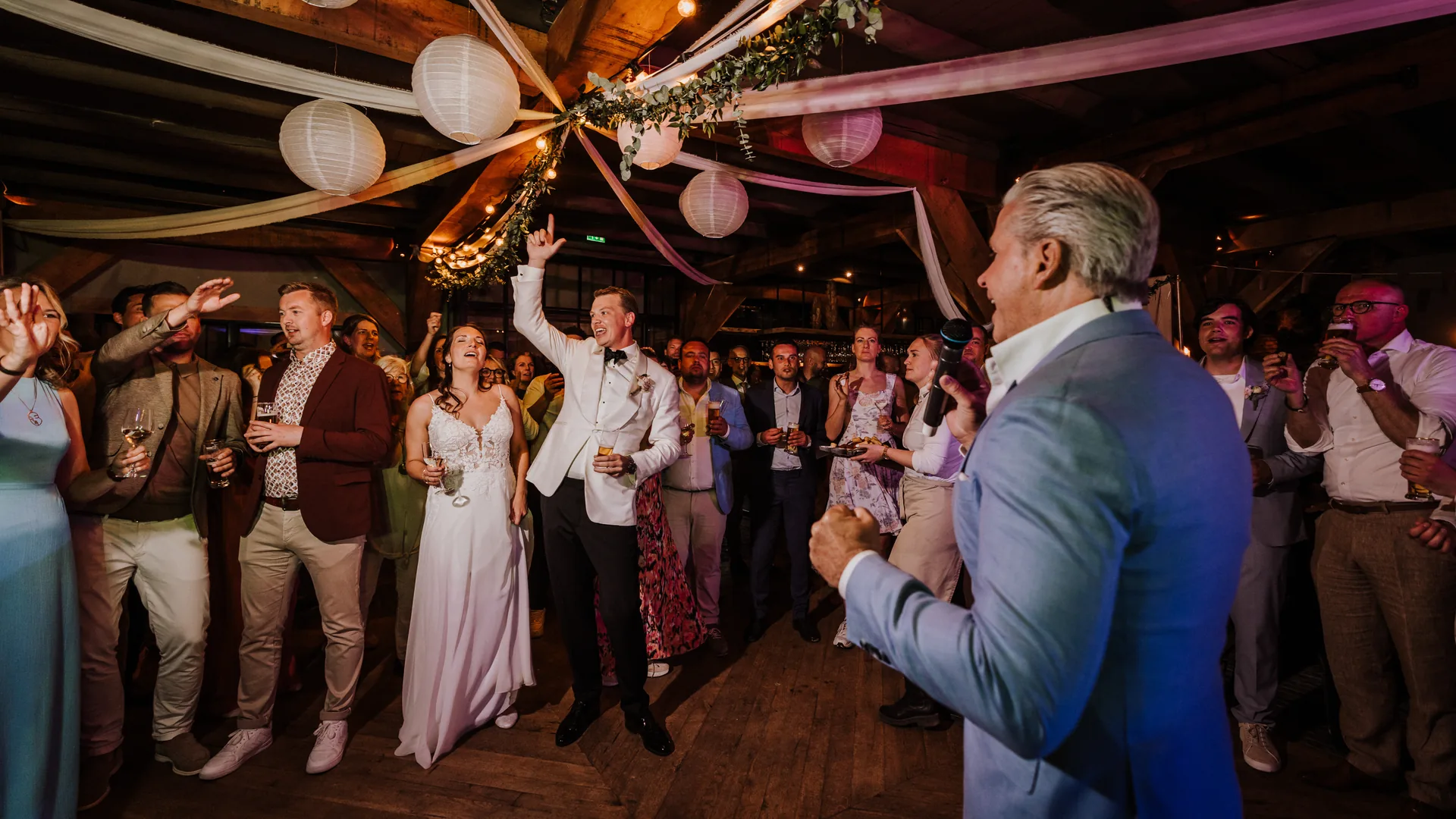 Feest in de Grote Zaal: onder de donkere eiken spanten hangen witte en roze lampionnen, banen stof en een groene slinger met lichtjes. Rechts zingt een man met grijs haar in een lichtblauw colbert in een microfoon; midden op de houten dansvloer juicht het bruidspaar mee, hij in een wit jasje met een arm in de lucht, zij in een witte kanten jurk met een glas in de hand. Rondom staan gasten met glazen, een paar steken hun handen op.