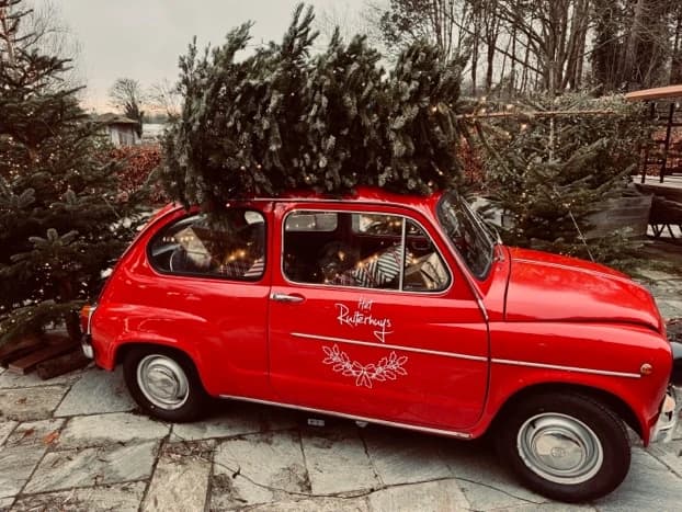 Een rode Fiat 600 met een kerstboom op het dak en "Het Ruiterhuys" in witte letters op de deur, geparkeerd op de natuurstenen vloer tussen kerstbomen met lichtjes.