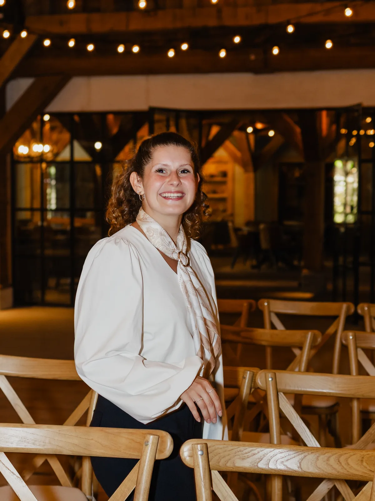 Alyssia staat lachend tussen de houten kruisstoelen van de ceremonie in de schuur, in een witte blouse met een zijden sjaal met ring, met lichtsnoeren en de glazen wand naar het restaurant achter haar.